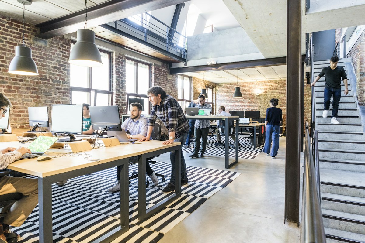 Équipe au travail dans un open space aux murs de briques, un collègue penché sur l'écran d'un autre (photo d'illustration)