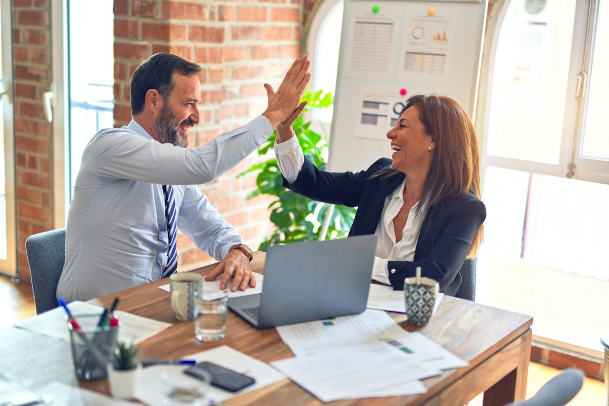 Un homme et une femme se tapant dans la main devant un ordinateur portable, documents sur la table (photo d'illustration)